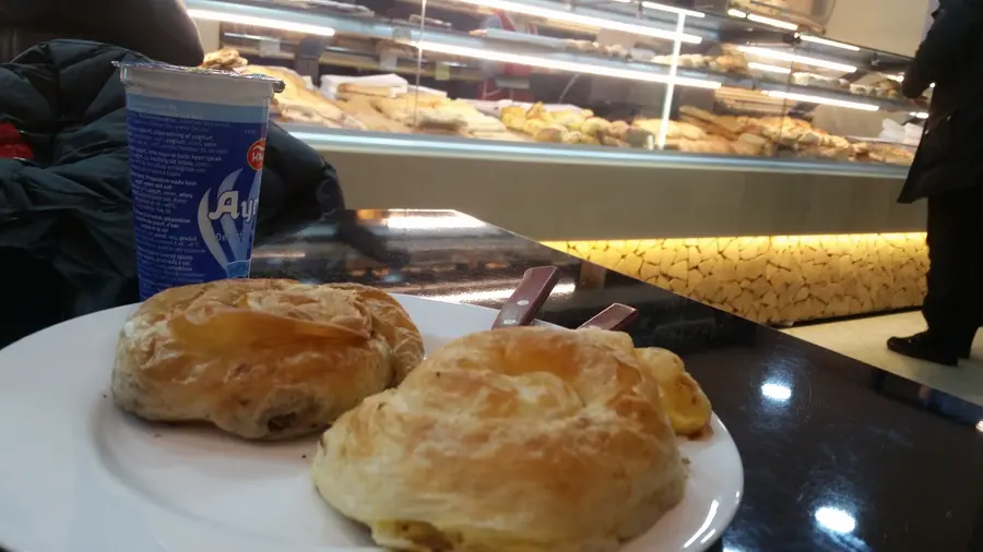 Zwei gerollte Burek auf einem Teller, dahinter ein Ayran und die beleuchtete Vitrine