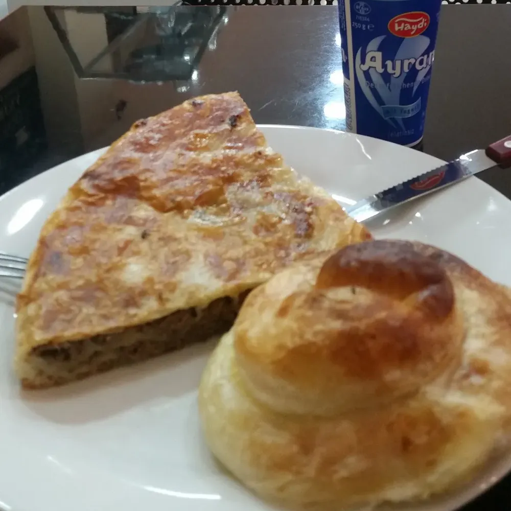 Burek mit Fleischfüllung und ein gerollter Burek auf einem Teller, dahinter ein Ayran