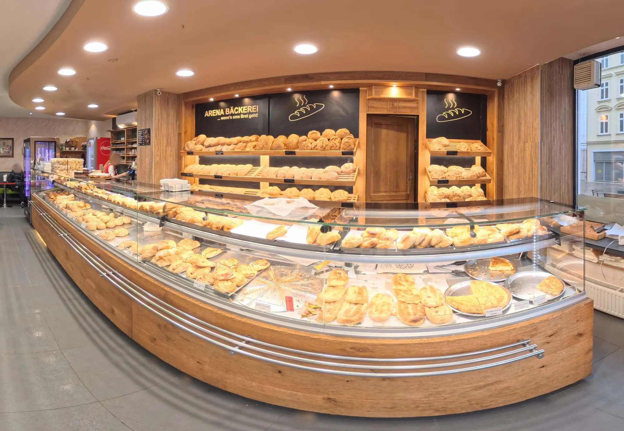 Die geschwungene Holztheke der Bäckerei Arena mit Brot, Burek und Gebäck, darüber die Tafel „Arena Bäckerei … wenn’s ums Brot geht!“
